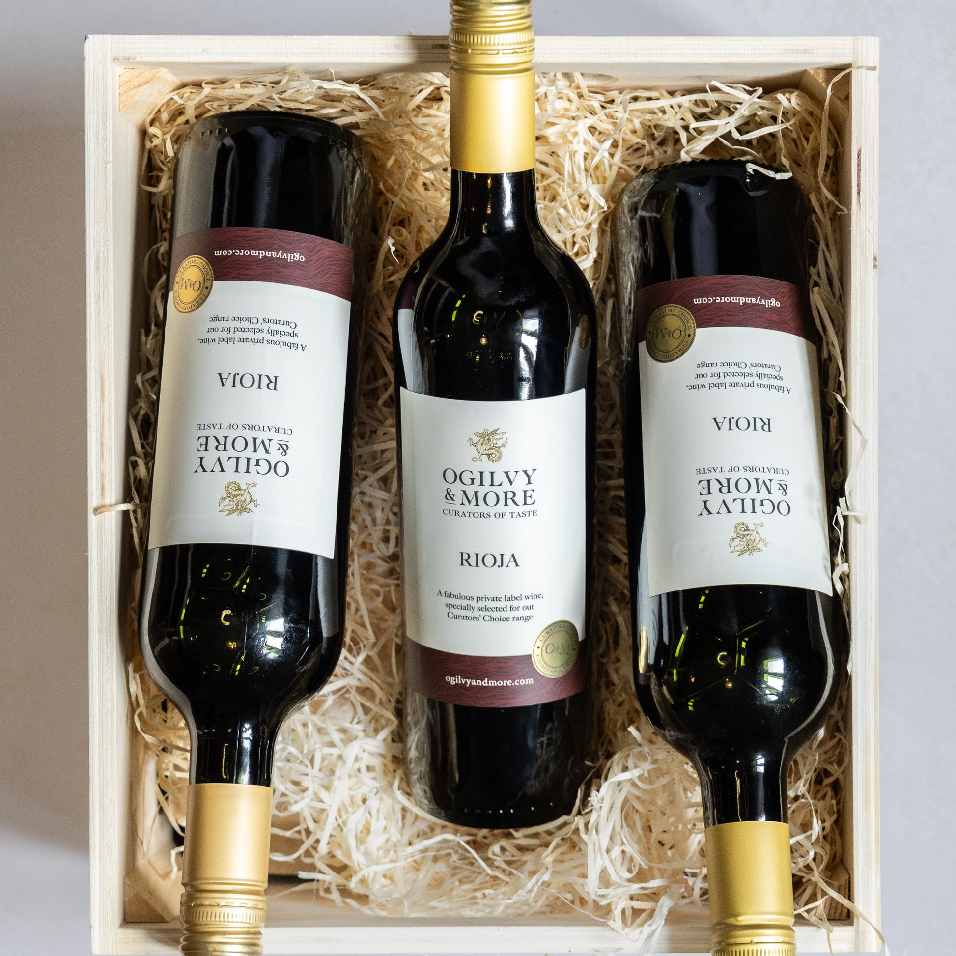 Three bottles of Ogilvy & More Rioja wine in a wooden gift box on a light gray background
