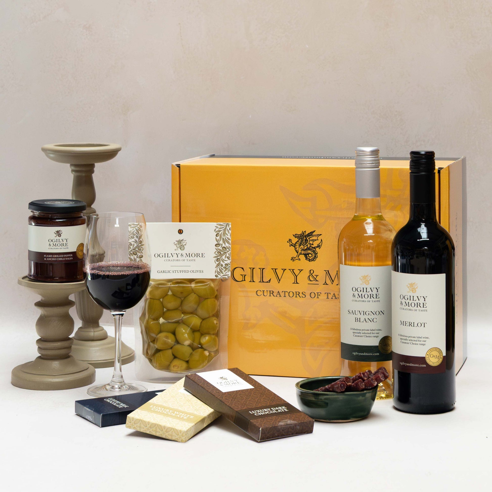 Ogilvy & More Wine gift set with bottles, a glass of wine, and snacks on a white surface.