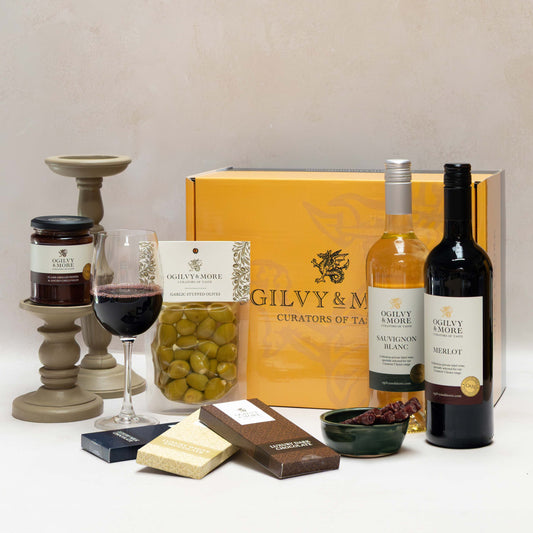 Ogilvy & More Wine gift set with bottles, a glass of wine, and snacks on a white surface.