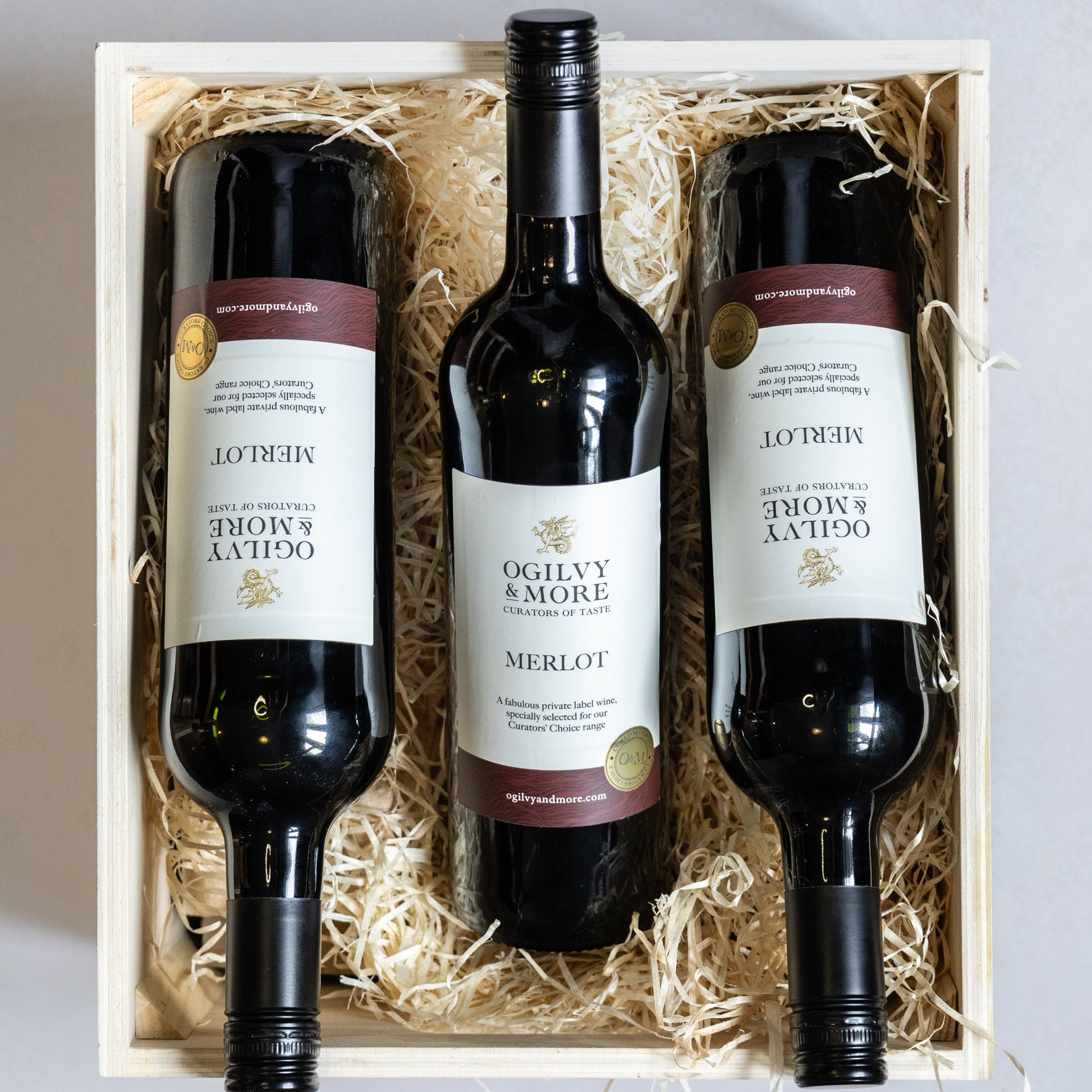 Three bottles of Ogilvy & More Merlot wine in a wooden gift box with straw interior.