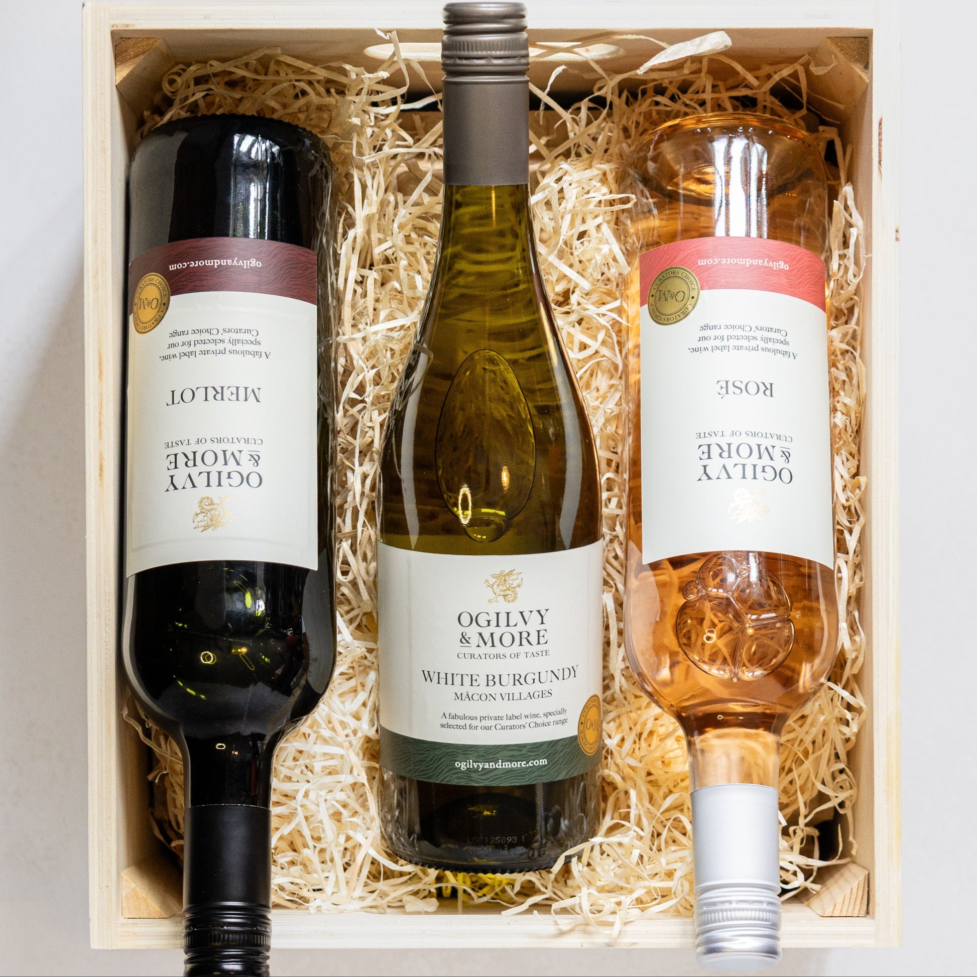 Three bottles of Ogilvy & More wine in a wooden box with straw lining on a white background