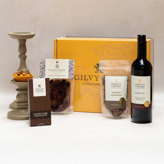 Ogilvy & More Dark & Delicious gift set with wine, snacks, and a candle on a white surface.