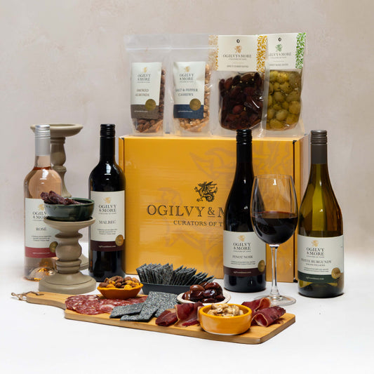 Ogilvy & More Wine bottles, charcuterie board, and snacks on a white surface with an Ogilvy & Armstrong box in the background.
